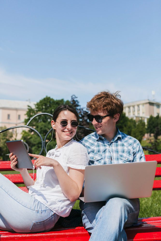 Two students sitting on a campus lawn with a laptop and tablet