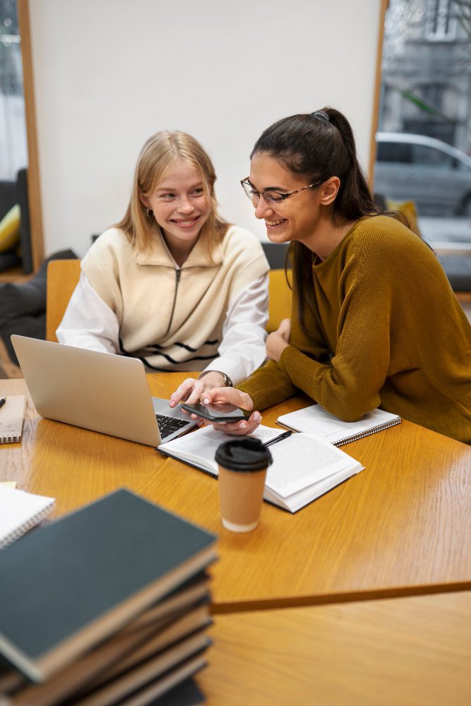 Two students working together on a laptop with textbooks on the table