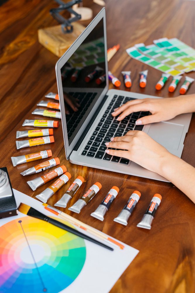 Website design - hands typing on a laptop surrounded by paint tubes and a colour wheel